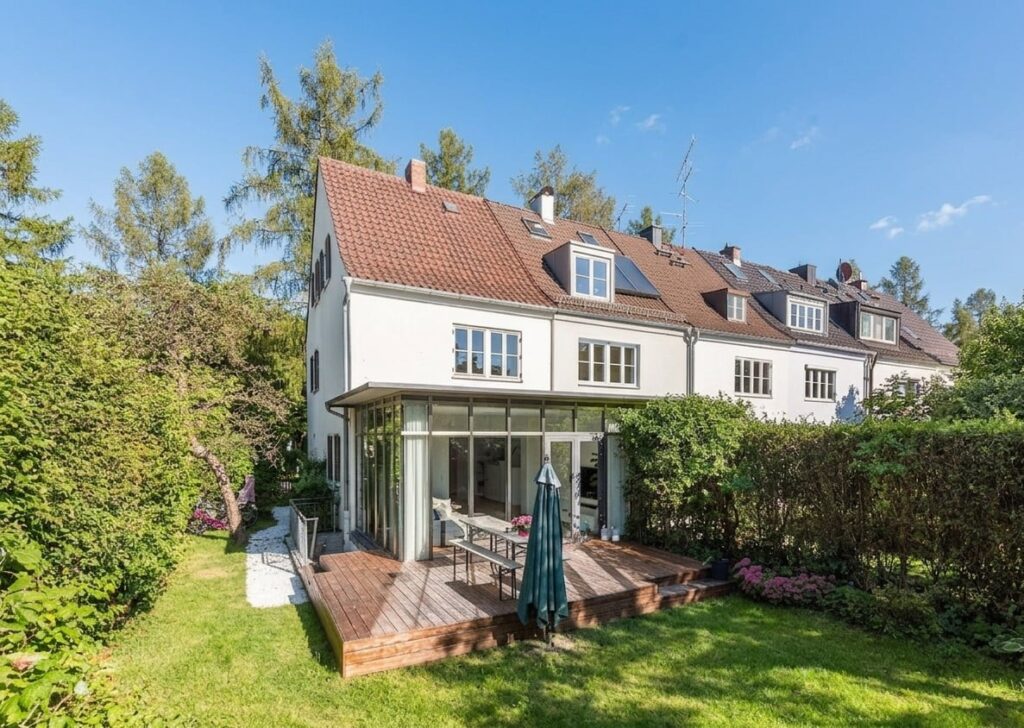 Reihenhaus mit rotem Ziegeldach, Wintergarten und Holzterrasse in einem grünen Garten mit Hecke und Bäumen im Hintergrund