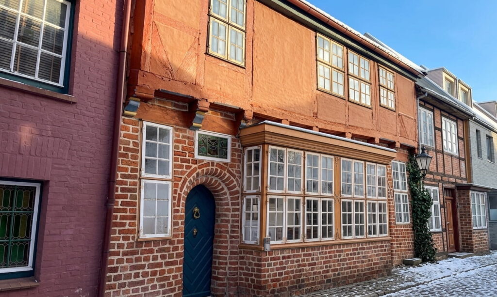 Fachwerkhaus mit roten Ziegeln, blauen Tür und mehreren Fenstern in einer Altstadtstraße mit leicht verschneitem Gehweg