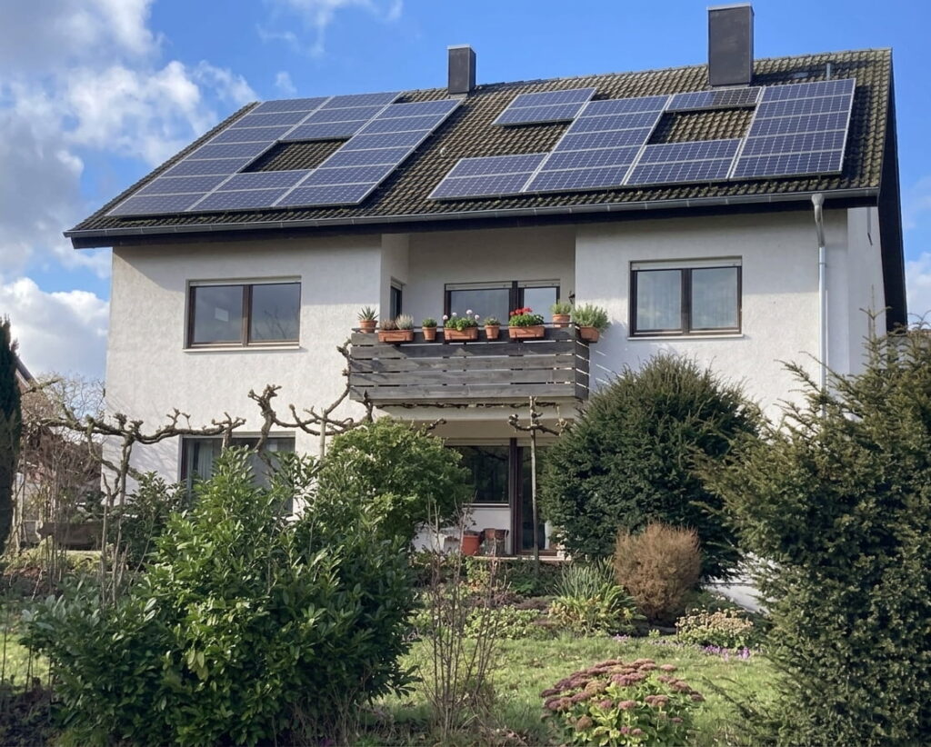 Zweistöckiges Wohnhaus mit Solarpaneelen auf dem Satteldach und Balkon mit Blumentöpfen, umgeben von Garten mit Sträuchern und Bäumen