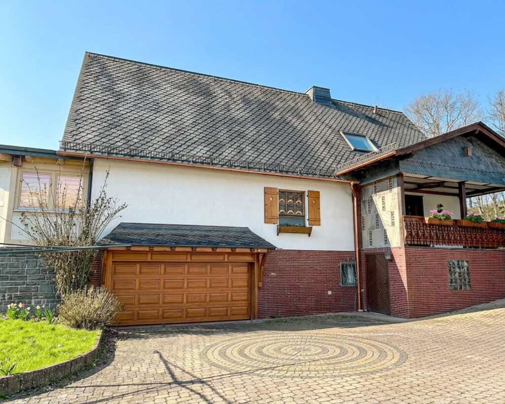 Haus mit Satteldach, Garage mit brauner Holztür, Fenster mit Holzläden und überdachtem Eingang mit Blumendekoration