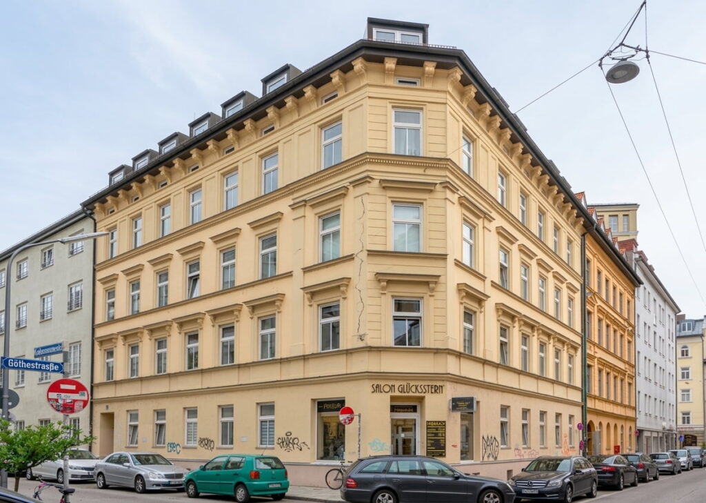 Mehrstöckiges Eckgebäude mit hellgelber Fassade und mehreren Fenstern in einer städtischen Straße mit parkenden Autos und Straßenschildern