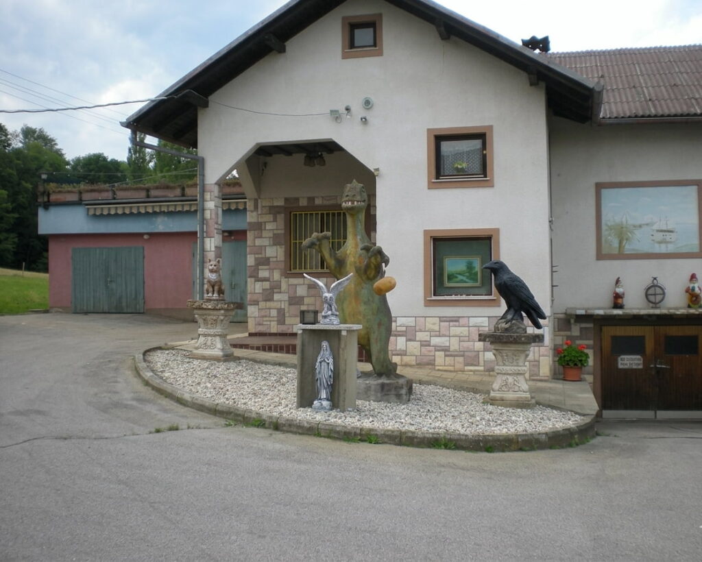 Haus mit weißer Fassade und braunem Dach, davor mehrere Skulpturen auf Kiesfläche, darunter eine große Figur, ein Adler und ein Rabe, im Hintergrund ein rotes Garagentor