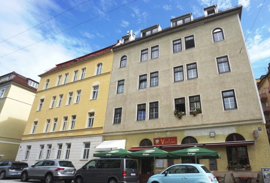 Mehrstöckiges Wohngebäude mit gelber und grauer Fassade, mehreren Fenstern und Straßencafé mit grünen Sonnenschirmen davor