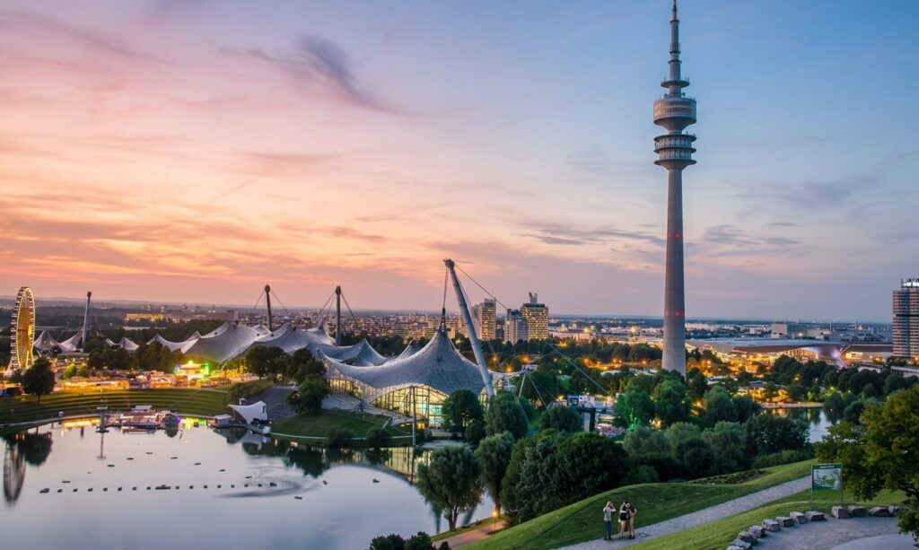 Blick auf den Olympiapark München mit dem Olympiaturm, dem Zeltdach des Olympiastadions, einem See und einer Stadtlandschaft im Abendlicht