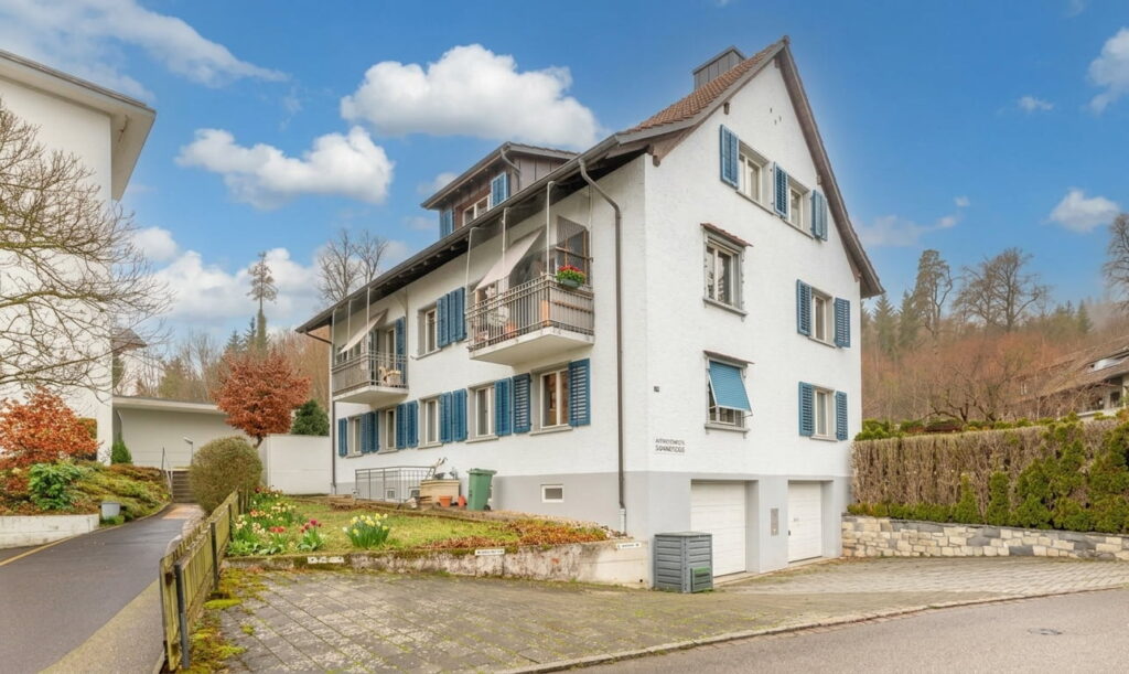Zweistöckiges Wohnhaus mit weißen Wänden, blauen Fensterläden und Balkonen in einer ruhigen Wohngegend mit Garten und gepflastertem Einfahrtsbereich
