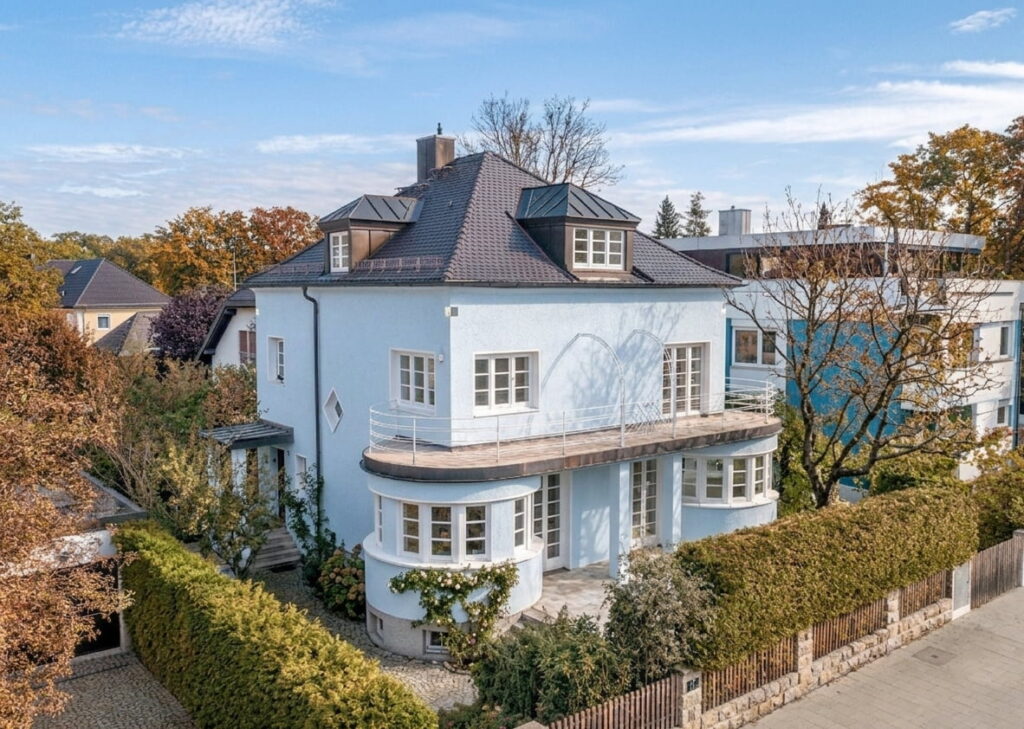 Großes weißes Eckhaus mit dunklem Walmdach, mehreren Fenstern und umlaufendem Balkon, umgeben von Hecken und Bäumen in einem Wohngebiet in Nymphenburg