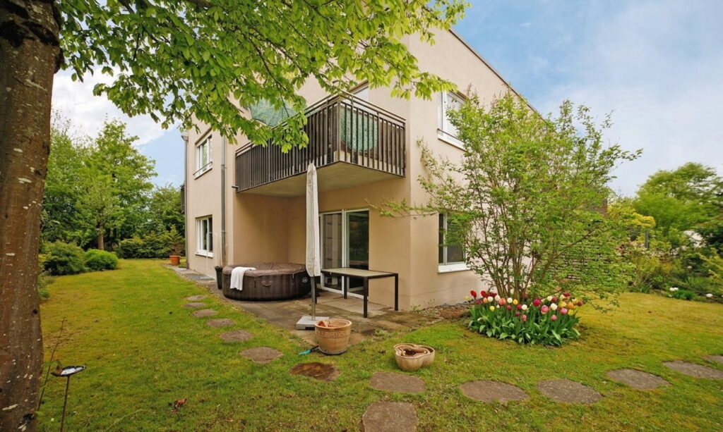 Eckansicht eines zweistöckigen Hauses mit Balkon, Garten mit Rasen, Blumenbeet und gepflastertem Weg