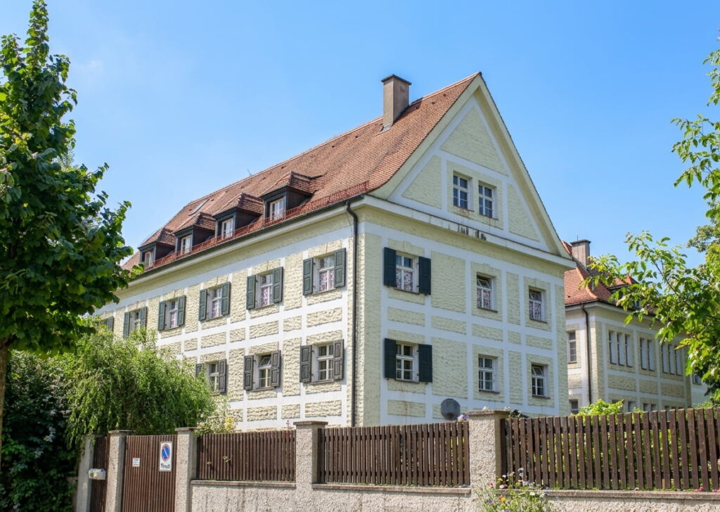 Großes, mehrstöckiges Wohnhaus mit Satteldach und Fensterläden hinter einem Zaun in Schwabing
