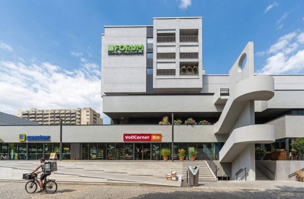 Modernes mehrstöckiges Gebäude mit Außenrampe und Geschäften im Erdgeschoss, darunter ein Bio-Laden mit rotem Schild 'VollCorner Bio' und ein Geschäft mit blauem Schild 'E center'