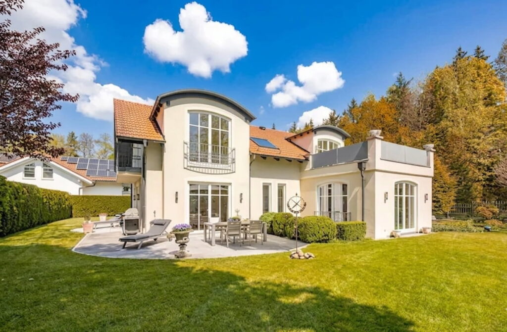 Großes modernes Einfamilienhaus mit zwei halbrunden Dachgauben, Terrasse mit Gartenmöbeln und Liegestühlen, umgeben von Rasen und Bäumen