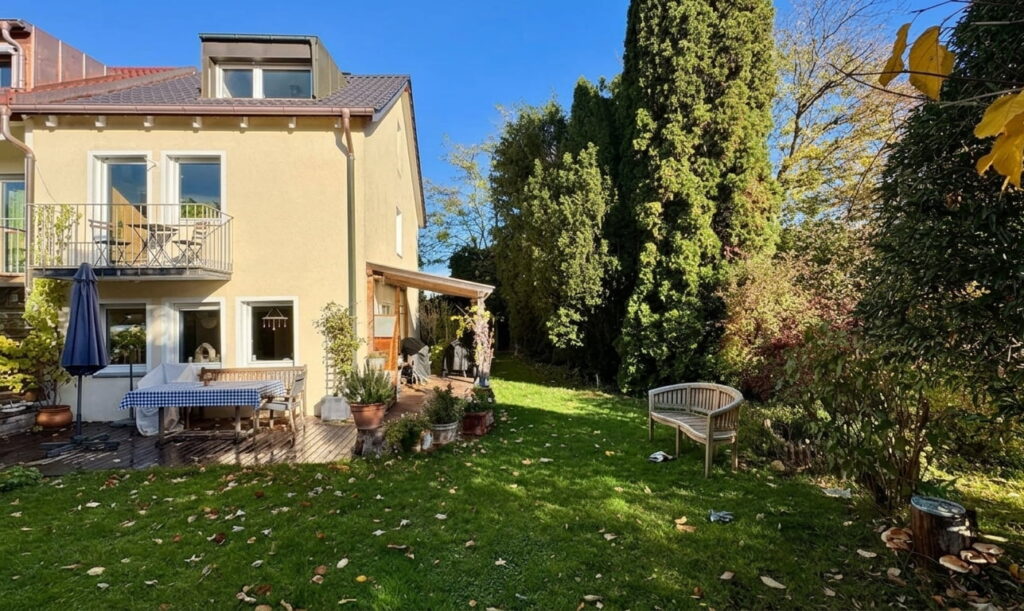 Zweistöckiges Haus mit Balkon und Terrasse neben einem gepflegten Garten mit Rasen, Büschen und einer Holzbank