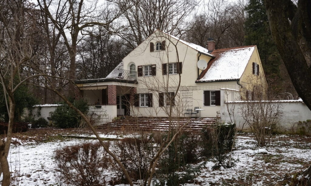 Zweistöckiges Haus mit Satteldach und weißen Außenwänden in winterlicher Umgebung mit leichtem Schneeboden und kahlen Bäumen