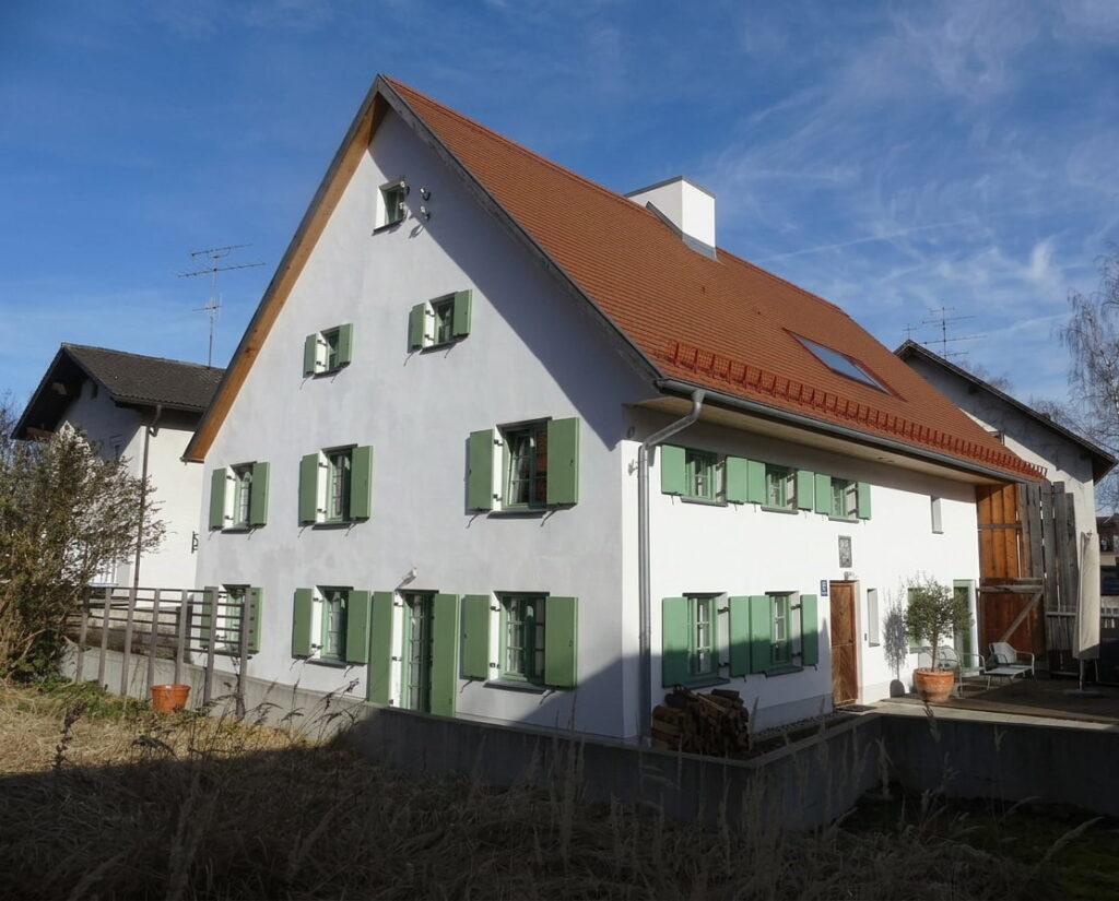 Großes, weißes Wohnhaus mit grün lackierten Fensterläden und rotem Satteldach in einem Wohngebiet.