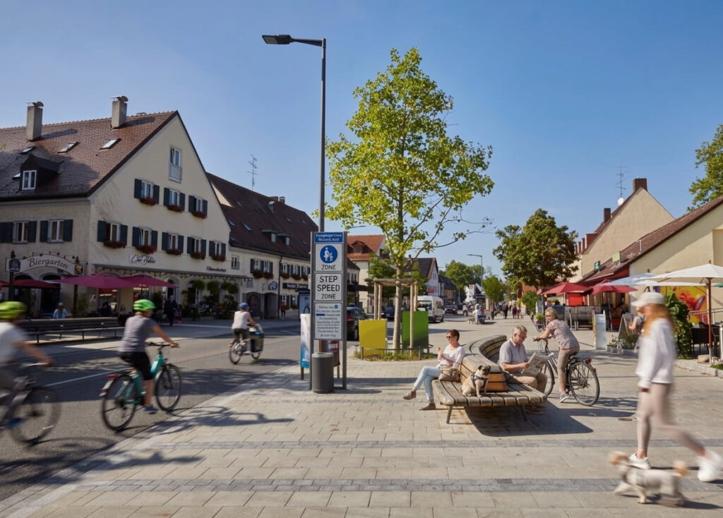 Belebter Platz in Trudering mit Radfahrern, Fußgängern, Sitzbänken und traditionellen Gebäuden mit Gasthäusern