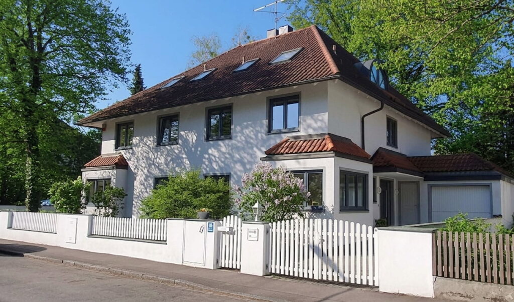 Zweistöckiges Einfamilienhaus mit Satteldach, weißen Fassaden, mehreren Fenstern und weißem Lattenzaun in einem grünen Wohngebiet