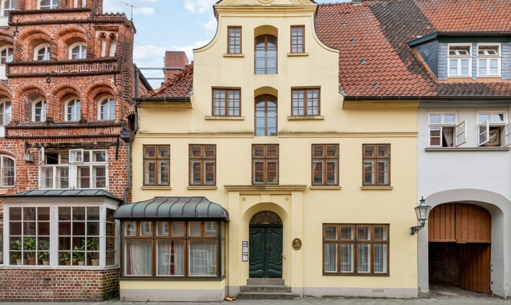 Fachwerkhaus mit gelber Fassade und grünem Eingang in einer Altstadtstraße neben einem Backsteinhaus mit Erker und einem weißen Gebäude mit Bogenfenster