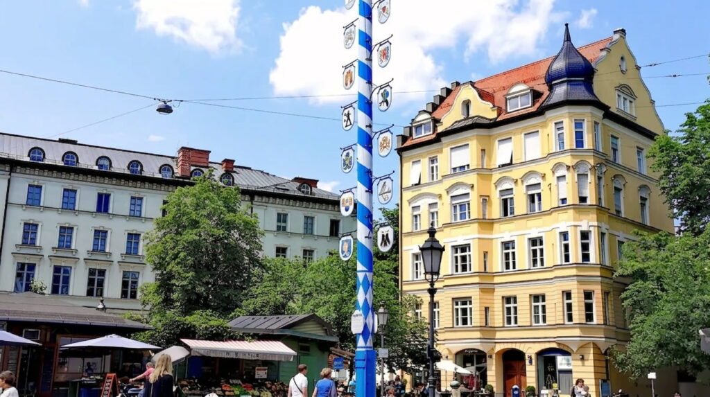 Marktplatz mit blau-weiß bemaltem Maibaum, umgeben von historischen mehrstöckigen Gebäuden und Menschen im Freien