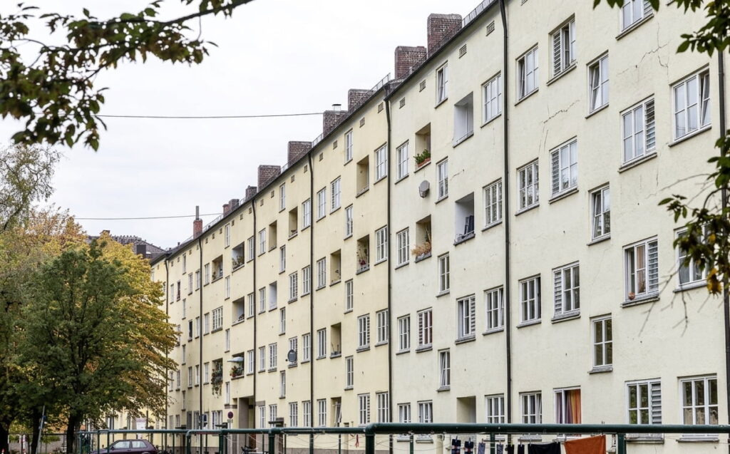 Mehrstöckiges Wohngebäude mit zahlreichen Fenstern und Balkonen in einer Reihe, davor Bäume und ein Zaun