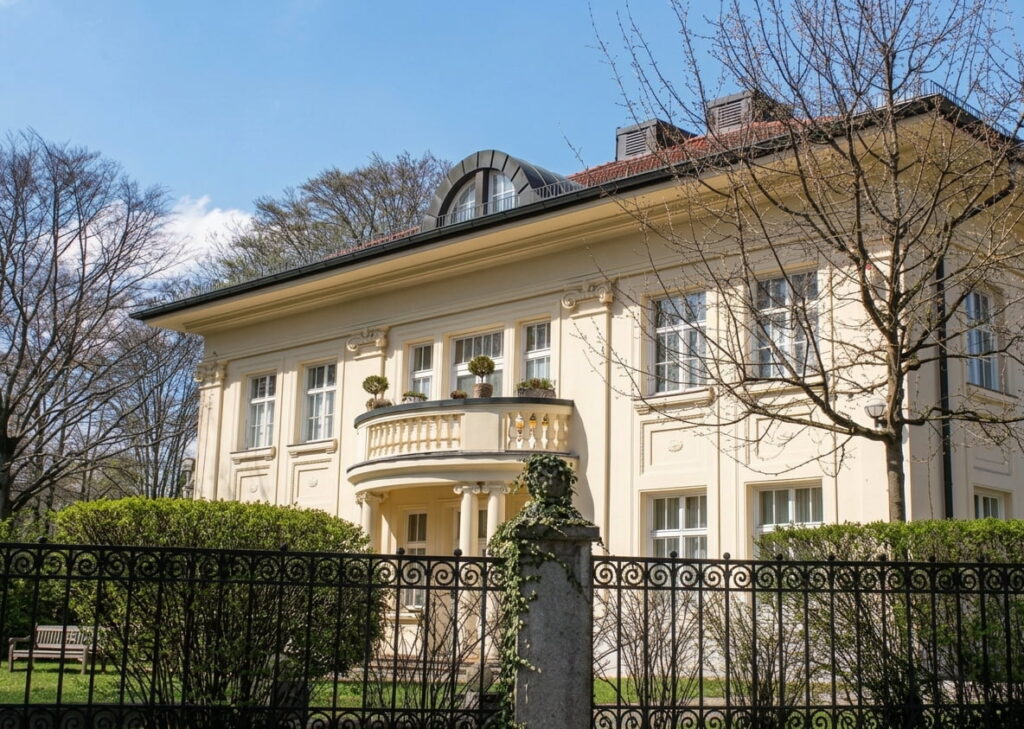 Großes klassizistisches Wohnhaus mit Balkon, Säulen und schmiedeeisernem Zaun im Vordergrund
