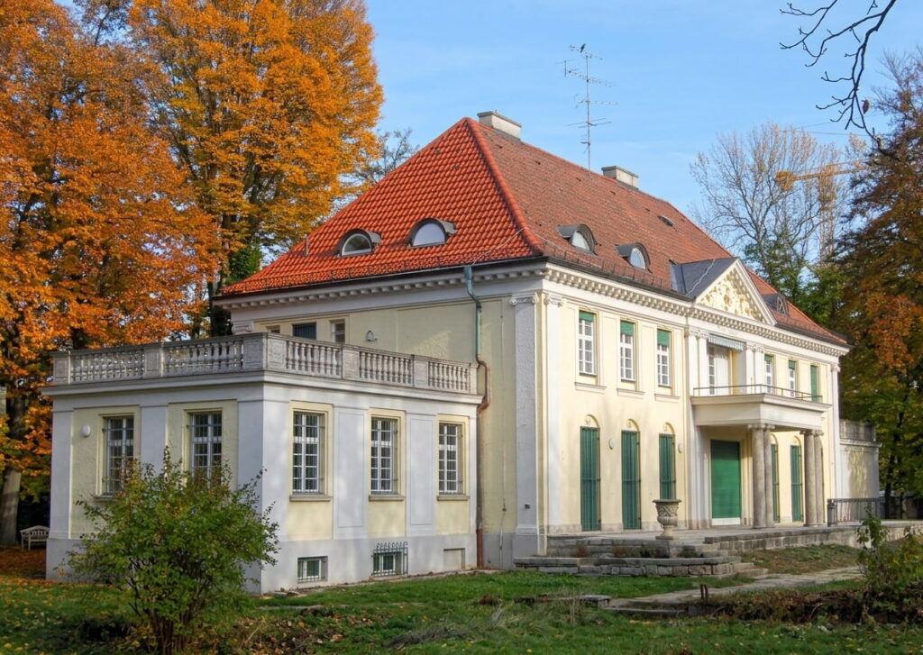 Großes klassizistisches Herrenhaus mit rotem Satteldach, grünen Fensterläden und umlaufender Terrasse, umgeben von herbstlich gefärbten Bäumen