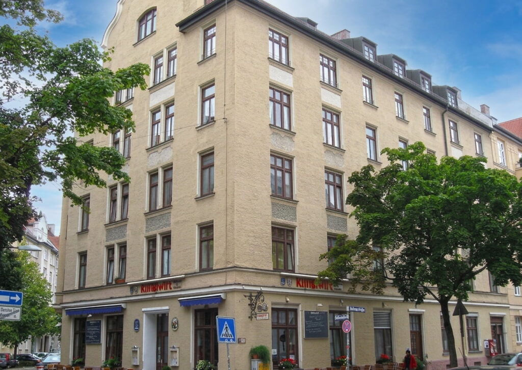 Mehrstöckiges Eckgebäude mit gelblicher Fassade, zahlreichen Fenstern und blauen Markisen an den Erdgeschossfenstern, umgeben von Bäumen und Straßenschildern in einem städtischen Viertel.