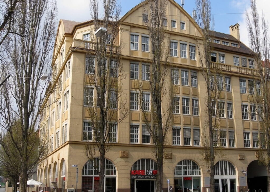Mehrstöckiges Eckgebäude mit zahlreichen Fenstern und Arkaden im Erdgeschoss, umgeben von kahlen Bäumen in einer städtischen Umgebung