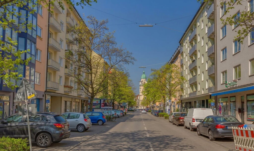 Straße in Maxvorstadt mit geparkten Autos und Bäumen, im Hintergrund eine Kirche mit grünem Turm und blauem Himmel