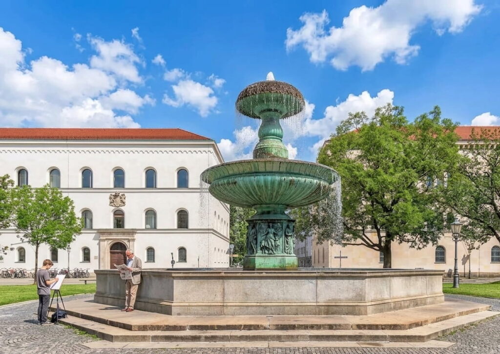 Große mehrstufige Brunnenanlage auf gepflastertem Platz vor historischen Gebäuden mit Bäumen und zwei Personen, eine malt und eine liest.