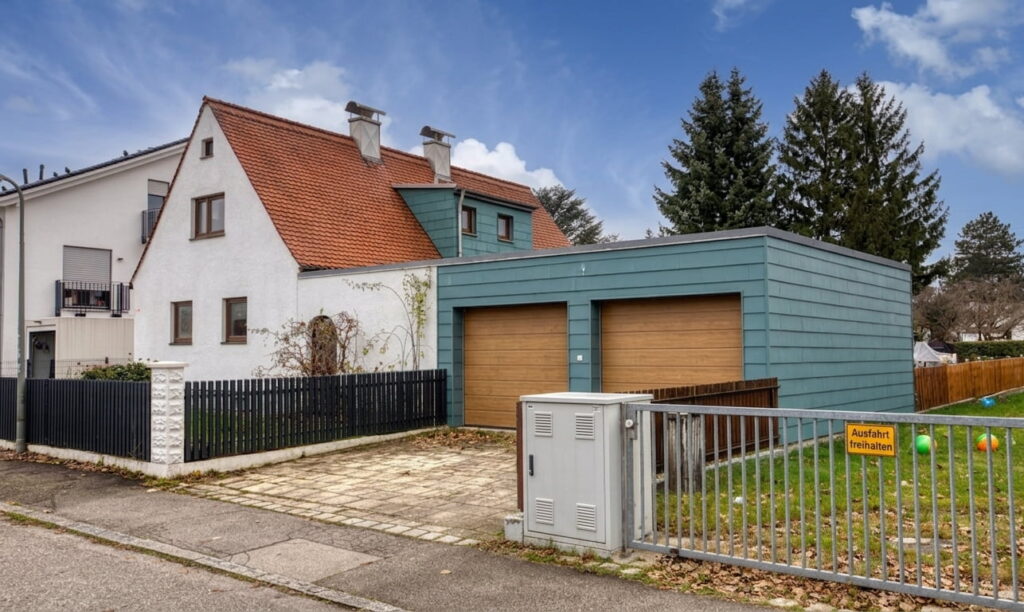 Zweistöckiges Wohnhaus mit rotem Satteldach neben modernem, flachem Doppelgaragengebäude mit blauer Verkleidung und Holztoren in Milbertshofen