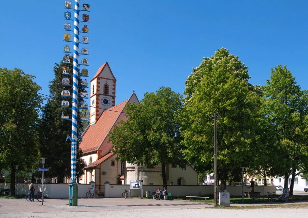 Kirche mit rotem Dach hinter Bäumen und blau-weiß gestreiftem Maibaum auf einem Platz in Moosach