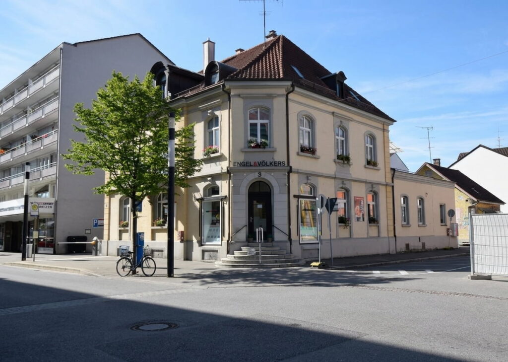 Eckgebäude mit klassischer Fassade und rotem Ziegeldach an einer Kreuzung mit Straßenschildern und einem Fahrrad vor einem Baum
