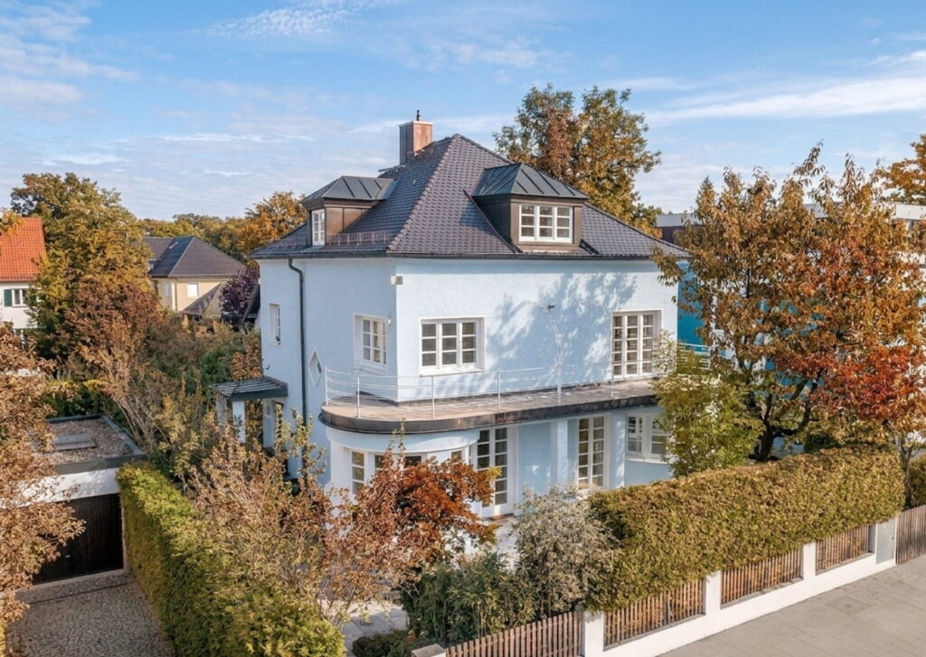 Großes, weißes Wohnhaus mit dunklem Walmdach umgeben von herbstlichen Bäumen und Hecken in einem Wohngebiet