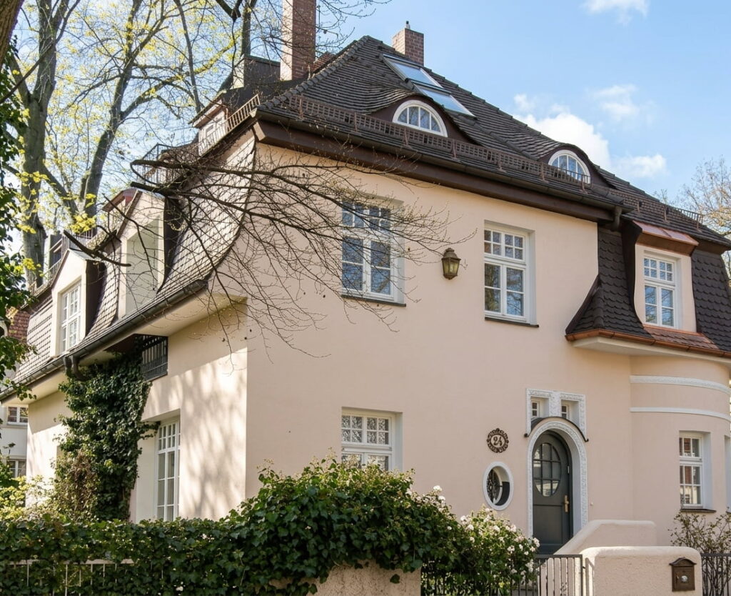 Großes, zweistöckiges Wohnhaus mit Satteldach, mehreren Fenstern und rundem Eingangstürfenster, umgeben von Büschen und Bäumen