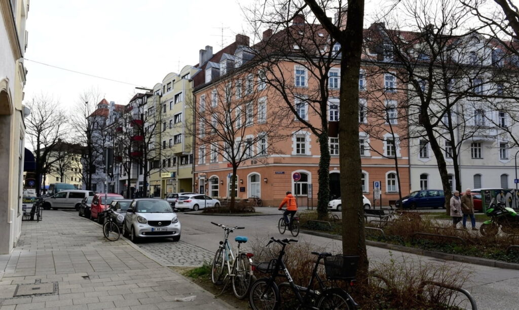 Straßenszene in Obergiesing mit mehrstöckigen Altbauten, geparkten Autos und Fahrrädern sowie einem Fußgänger in orangefarbener Jacke