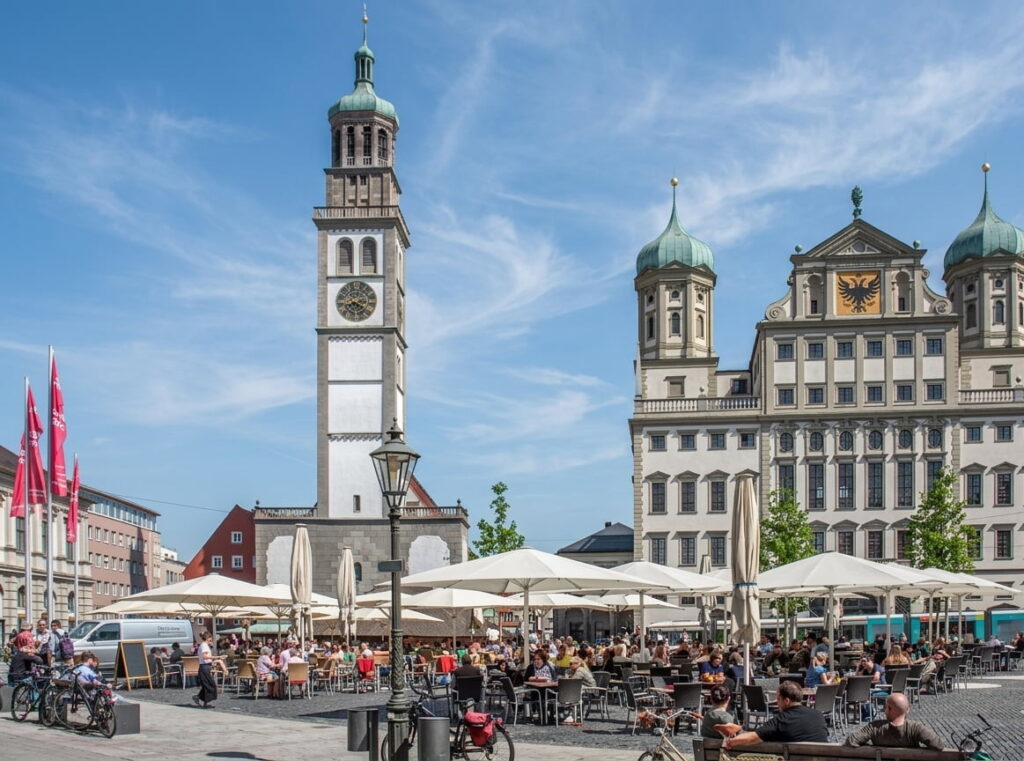 Großer belebter Platz mit vielen Tischen und Sonnenschirmen vor historischen Gebäuden und einem hohen Turm mit Uhr unter blauem Himmel