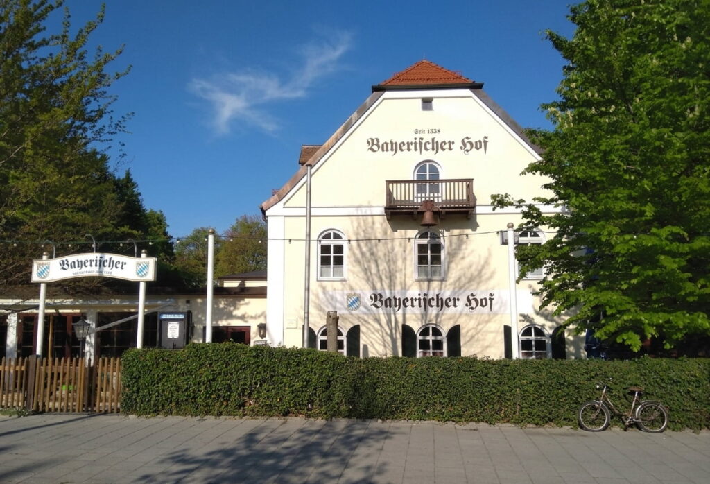 Fachwerkhaus mit Schriftzug 'Bayerischer Hof' und kleinem Balkon, umgeben von Bäumen und Hecke, davor Fahrrad an Gehweg
