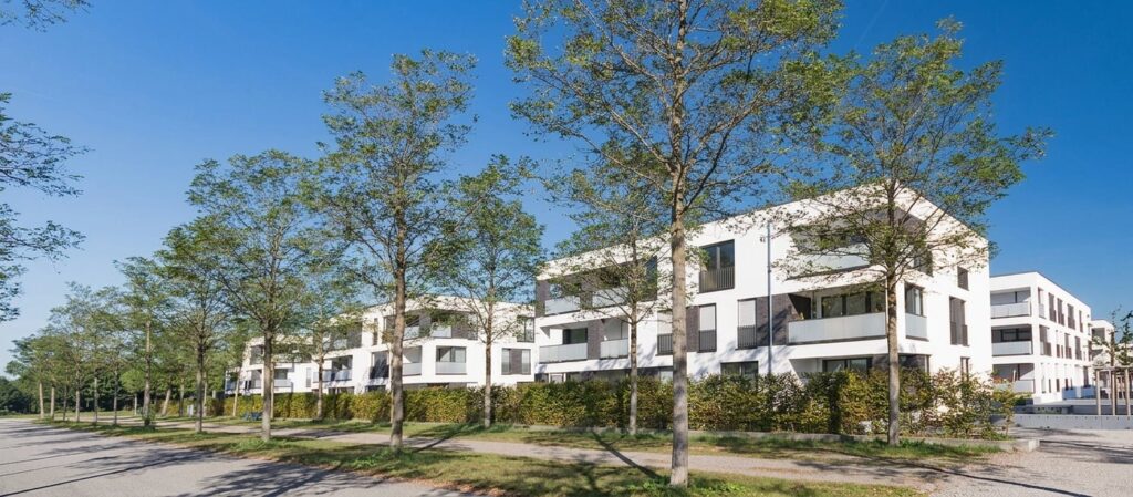 Mehrere moderne, weiße Wohngebäude mit Flachdächern und großen Fenstern, umgeben von Bäumen und einem Gehweg unter blauem Himmel