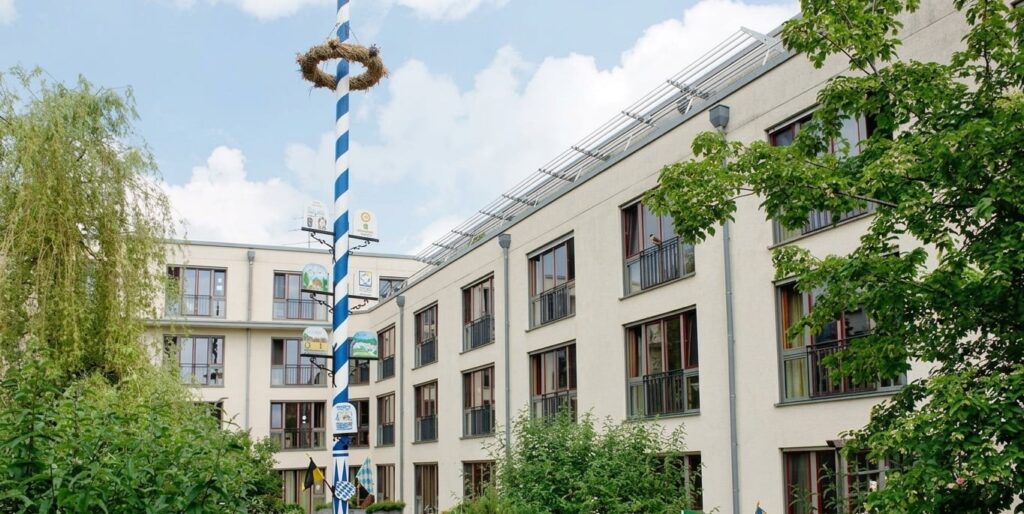 Mehrstöckiges Wohngebäude mit Balkonen und Bäumen im Vordergrund sowie einem traditionellen Maibaum mit blau-weißen Streifen