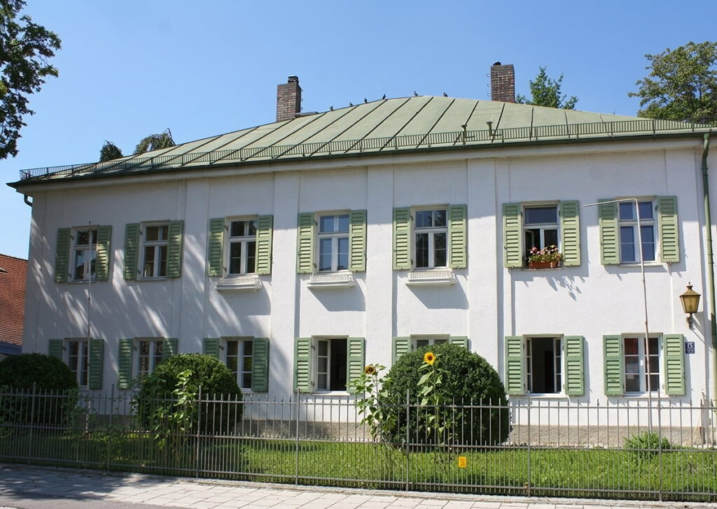 Zweigeschossiges Wohnhaus mit grünem Satteldach, grünen Fensterläden und Garten mit Sträuchern hinter einem Metallzaun