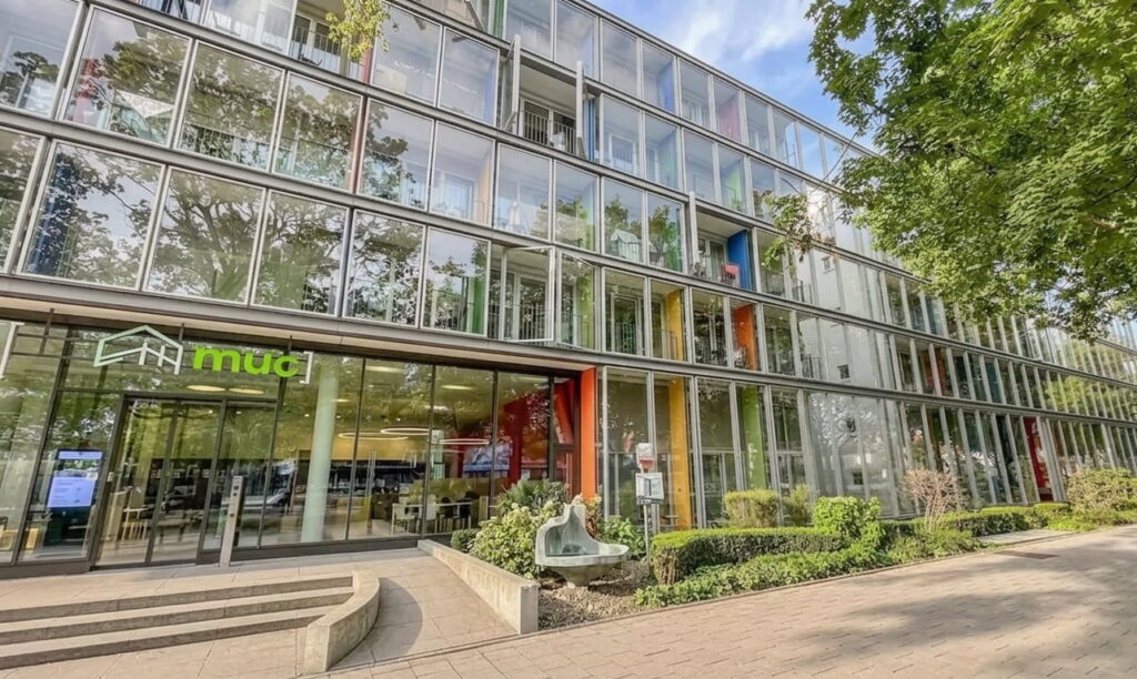 Moderne Glasfassade eines mehrstöckigen Bürogebäudes mit grünem Schriftzug 'mud' am Eingang und umgebender Bepflanzung