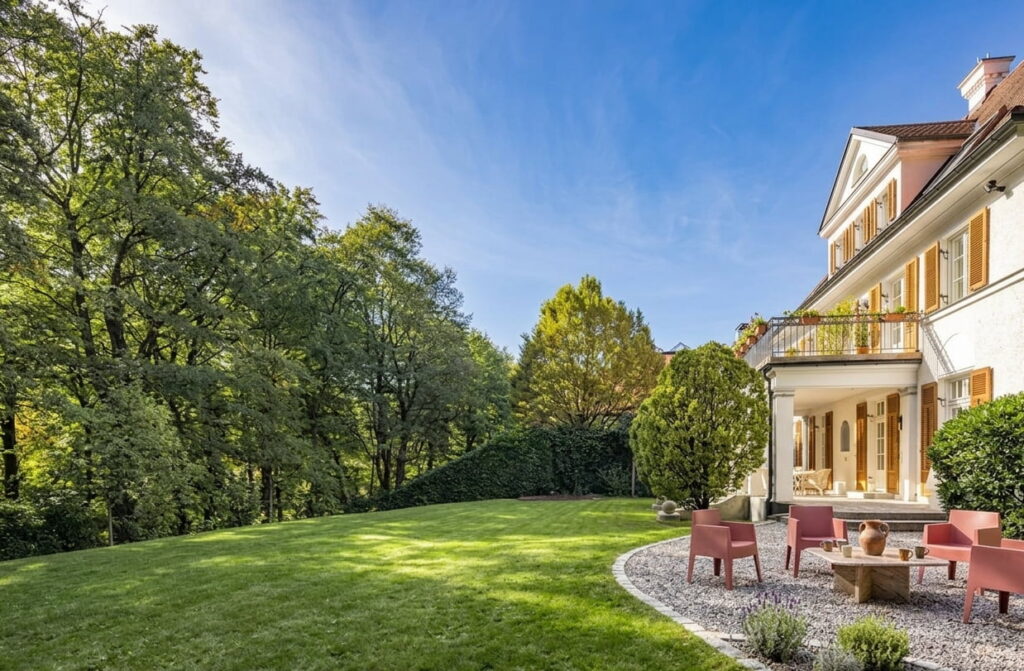 Großes, gepflegtes Grundstück mit Rasenfläche und Sitzbereich neben einem mehrstöckigen Haus mit Balkon und großen Fenstern