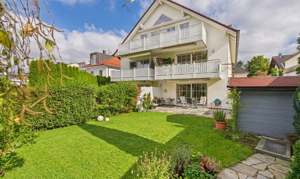 Zweistöckiges Wohnhaus mit Balkon und Terrasse, umgeben von gepflegtem Garten mit Rasen, Hecke und Blumenbeeten