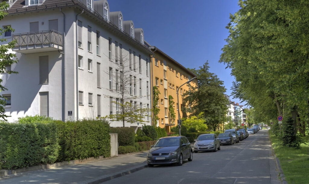 Straße mit parkenden Autos und mehrstöckigen Wohnhäusern auf der linken Seite sowie Bäumen auf der rechten Seite bei klarem Himmel