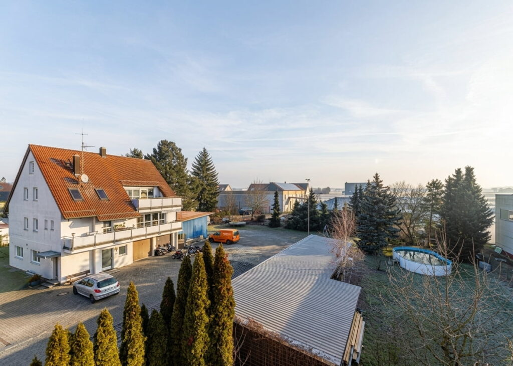 Mehrfamilienhaus mit rotem Satteldach, mehreren Balkonen und umliegendem Garten mit Bäumen und einem runden Pool, davor ein Parkplatz mit zwei Fahrzeugen