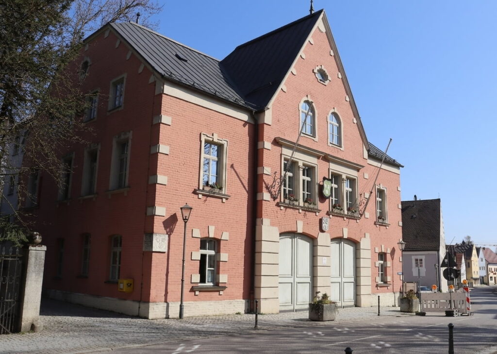 Zweistöckiges rotes Backsteingebäude mit spitzem Dach, mehreren Fenstern und zwei großen Toren an der Vorderseite, umgeben von einer gepflasterten Straße und einem Zaun.