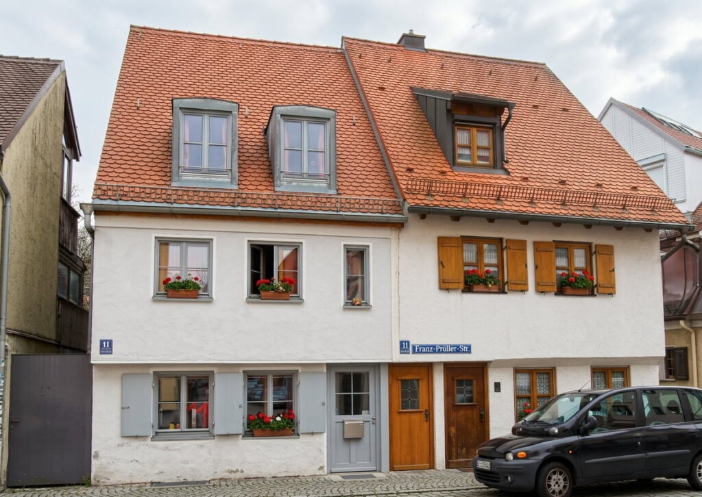 Zweistöckiges Wohnhaus mit weißer Fassade, roten Dachziegeln, mehreren Fenstern mit Blumenkästen und zwei Eingängen, davor ein schwarzes Auto auf Kopfsteinpflaster.