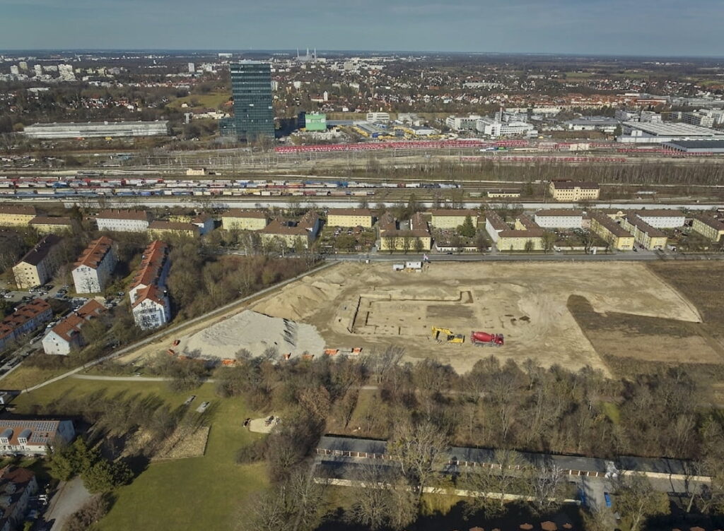 Luftaufnahme eines Baugrundstücks mit Baumaschinen im Vordergrund, dahinter Wohngebäude, eine Bahnstrecke und ein hoher Bürogebäudekomplex in Berg am Laim