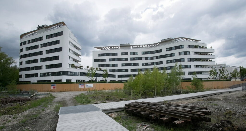 Mehrere moderne, weiße Wohngebäude mit abgerundeten Ecken vor bewölktem Himmel und Baustellenbereich im Vordergrund