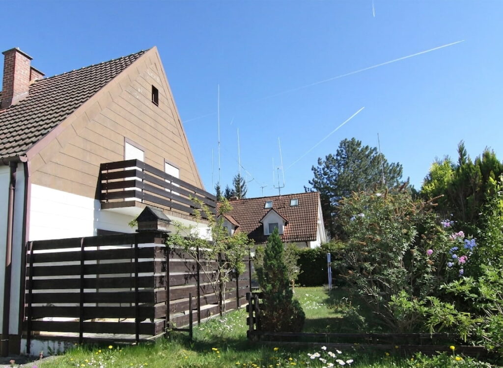 Garten mit Rasen, Blumen und Sträuchern neben einem Haus mit braunem Dach und weißer Fassade unter blauem Himmel
