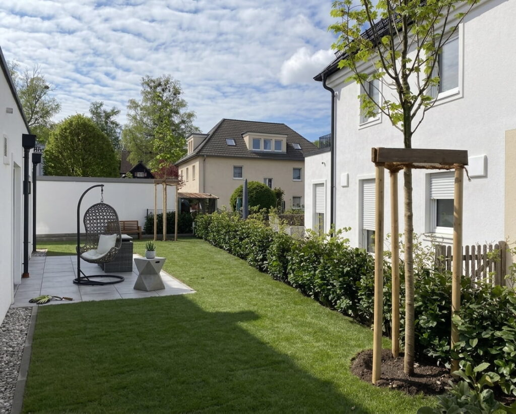 Moderner Garten mit gepflegtem Rasen, jungen Bäumen mit Stützpfosten, Hecke entlang eines weißen Hauses und einem hängenden Korbsessel auf einer Terrasse